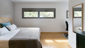 A bedroom with a bed, a desk, a chair, and a window with a view of greenery.