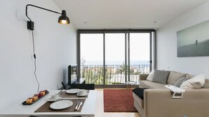 A modern living room with a dining area, a sofa, a TV, and a balcony with a view of the sea.