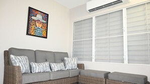 A grey sectional sofa with patterned cushions, a wicker accent chair, and a large window with blinds. - Jlo’s Place/Next to Airport/PrimeLocation/Fastnet (Carolina)