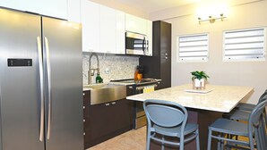 A modern kitchen with a stainless steel refrigerator, a microwave, and a sink. There are two bar stools and a small island with a plant. - Jlo’s Place/Next to Airport/PrimeLocation/Fastnet (Carolina)