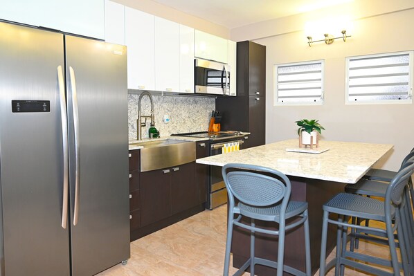 A modern kitchen with a stainless steel refrigerator, a microwave, and a sink. There are two bar stools and a small island with a plant. - Jlo’s Place/Next to Airport/PrimeLocation/Fastnet (Carolina)