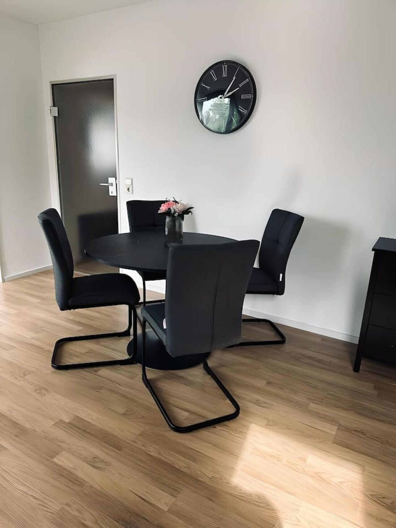 A modern dining area with a black table and chairs, a round wall clock, and a door in the background.