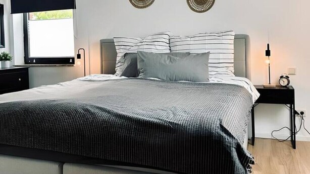 A modern bedroom with a large bed, a window with a blind, and decorative wall art. - FeWo Burgblick - Sun terrace & castle view (Bad Bentheim)