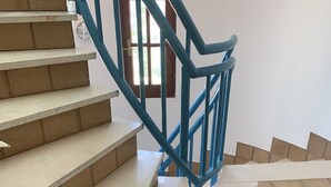 Een trap met blauwe leuningen, twee blauwe emmers op de overloop en een witte muur met een raam op de achtergrond.
