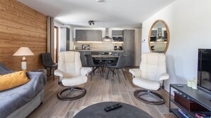 A modern living room with a sofa, two armchairs, a coffee table, and a TV stand. A kitchen with grey cabinets and a dining area with a table and chairs is visible in the background. - Le Bierge - Station de ski accessible à pied (Vars)