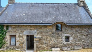 A stone building with a sloped roof, a small arched window, and a door with a sign above it.
