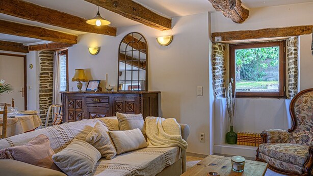 A cozy living room with a sofa, a wooden coffee table, and a chair. There is a fireplace with a mirror above it, and a window with a view of greenery outside.