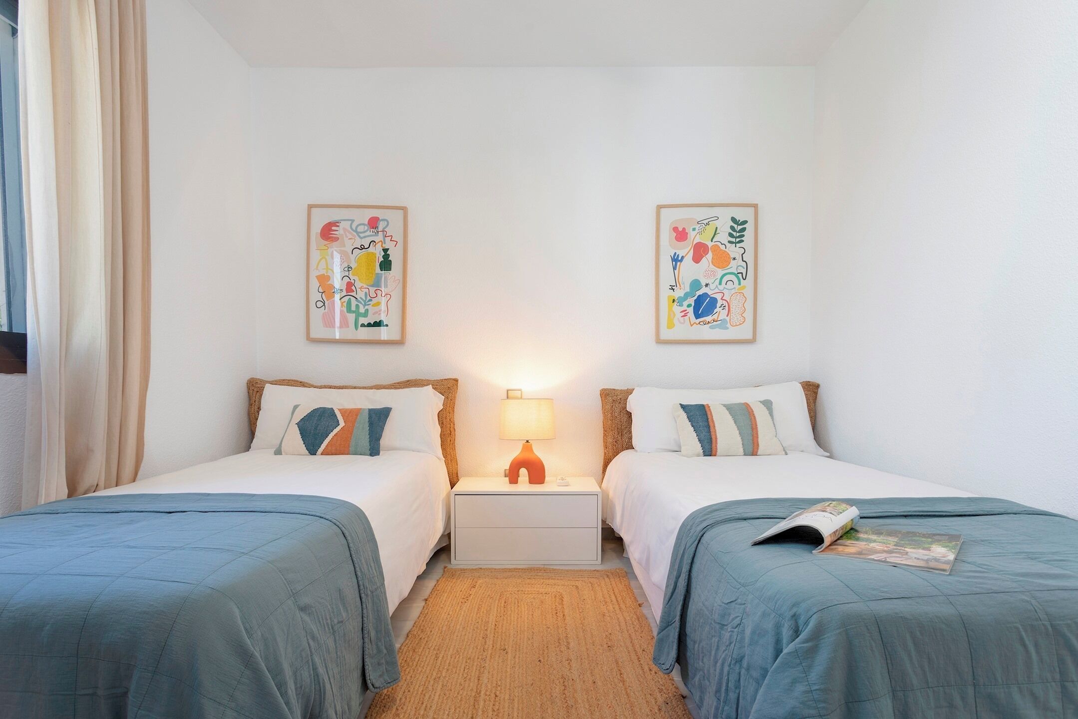 Quarto de hotel com duas camas, uma mesa de cabeceira com abajur e duas pinturas abstratas nas paredes.
