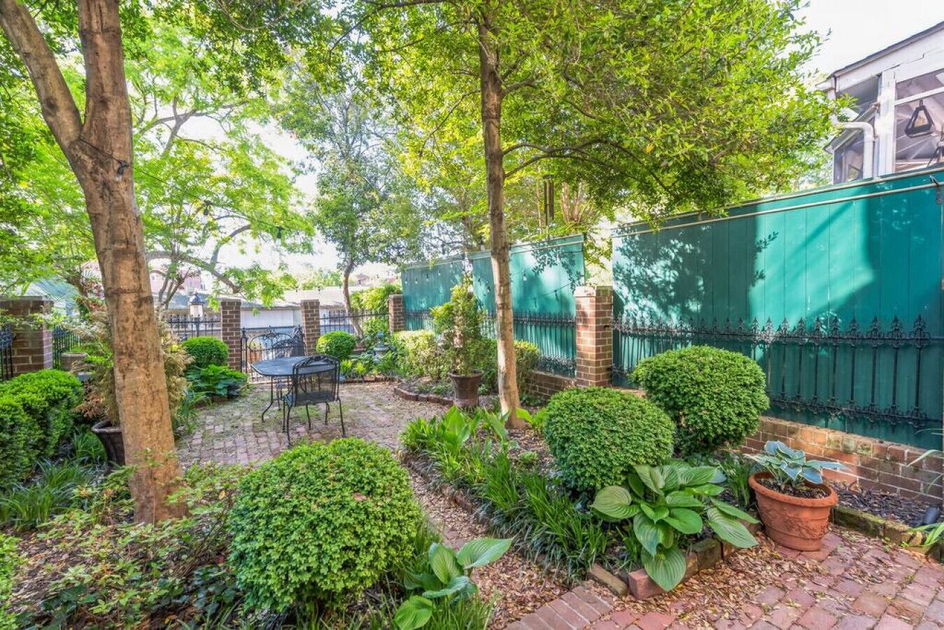 Un giardino con un patio in mattoni, un tavolo in metallo e una recinzione verde.