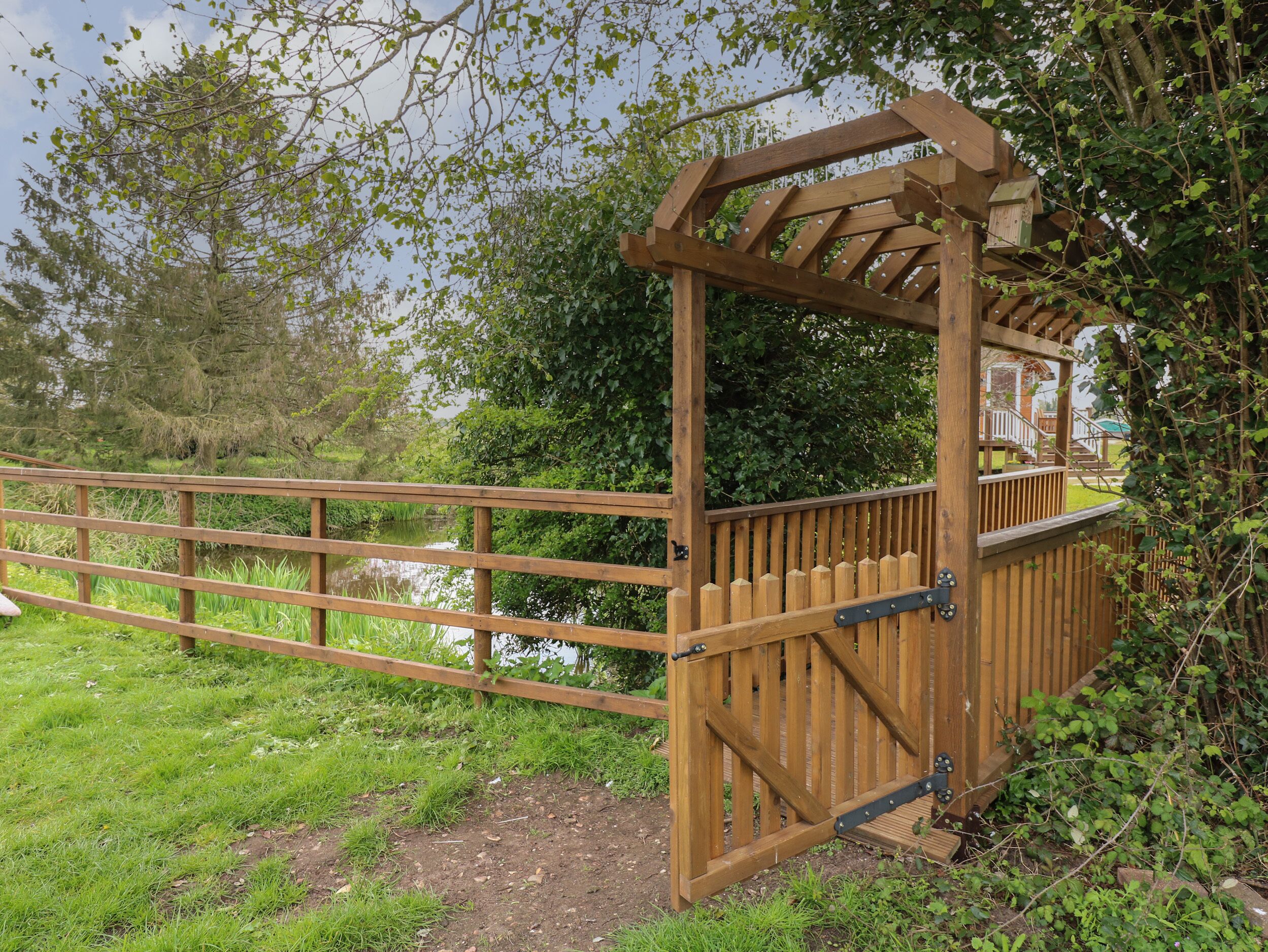 A wooden pergola with a metal gate, surrounded by greenery and a body of water.