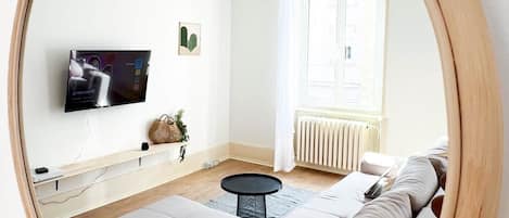 A modern living room with a large round mirror, a sofa, a coffee table, a TV, and a window with curtains.