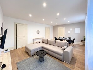 A modern living room with a sectional sofa, a dining table with chairs, a small round table with a remote control, and a grey rug.