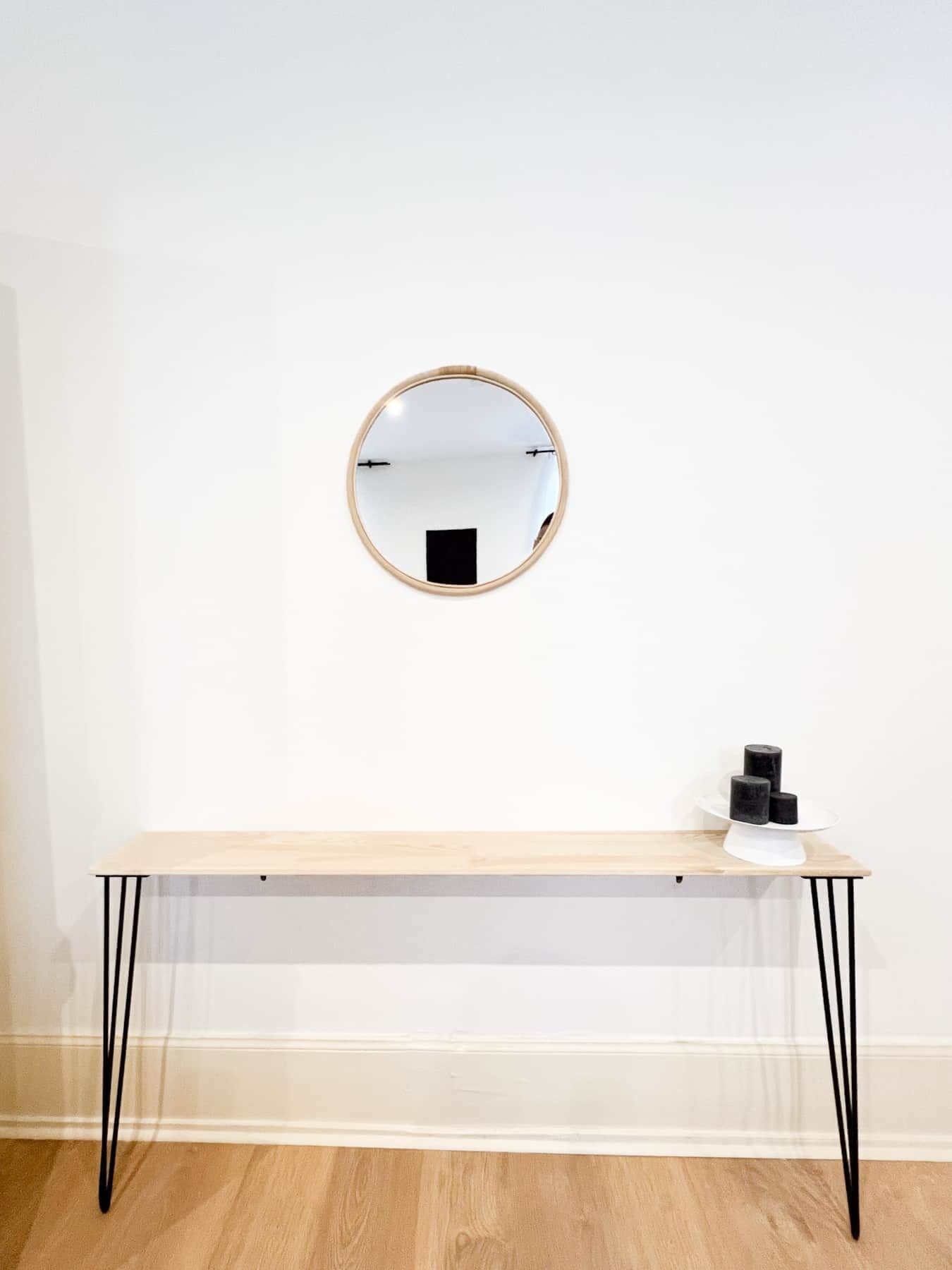 Une pièce au décor minimaliste, avec un miroir rond accroché au mur, une table en bois sur laquelle se trouve un objet noir, et une chaise blanche.