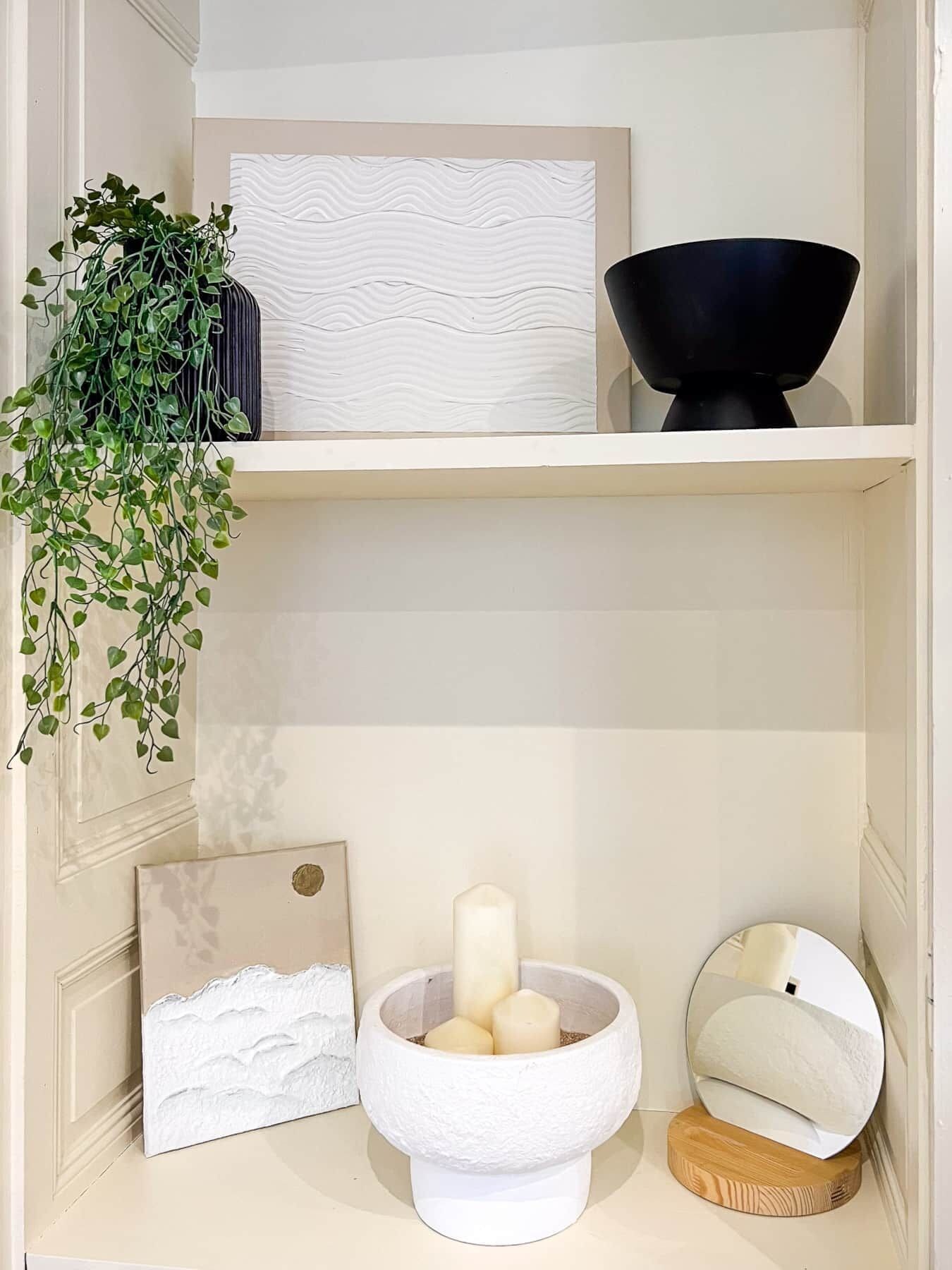 Een plank met wanddecoratie in witte textuur, een zwarte schaal, een witte kandelaar met kaarsen, een ingelijst beeld en een hangende plant.