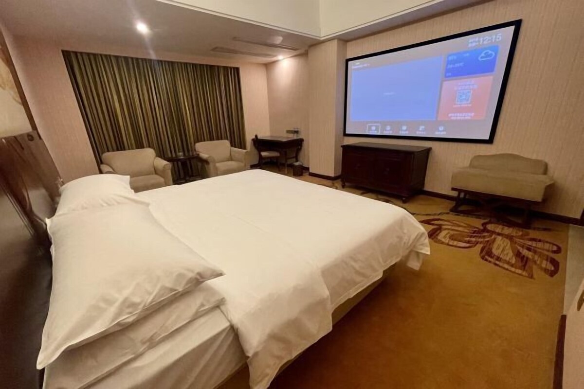 A hotel room with a large bed, a desk, two chairs, a TV screen, and a curtain.