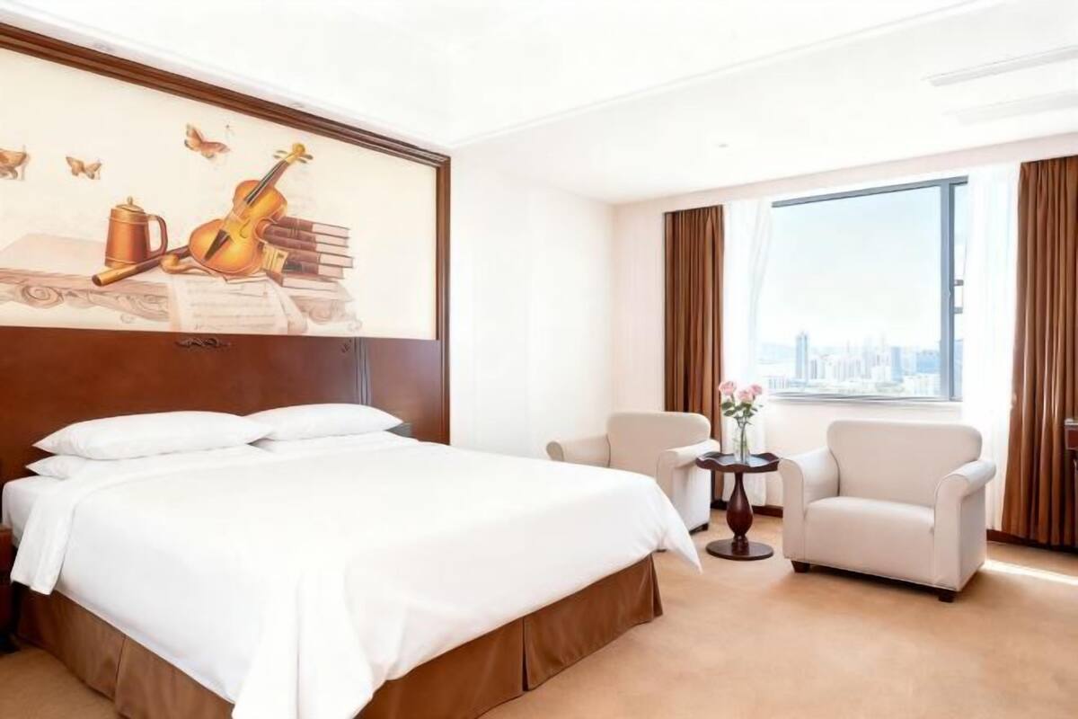 A hotel room with a large bed, a chair, a small table with flowers, and a city view.
