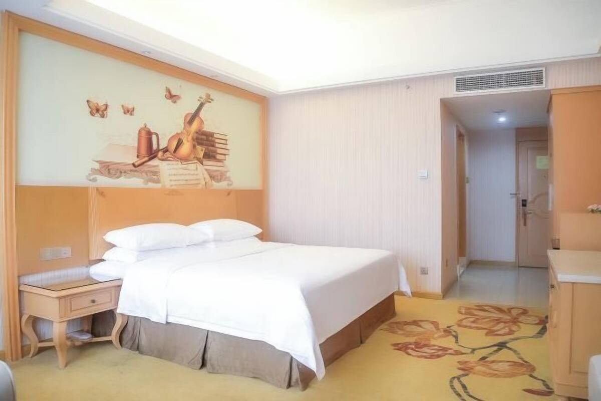 A hotel room with a bed, a nightstand, a wall with a mural, a door, and a carpet with a floral pattern.