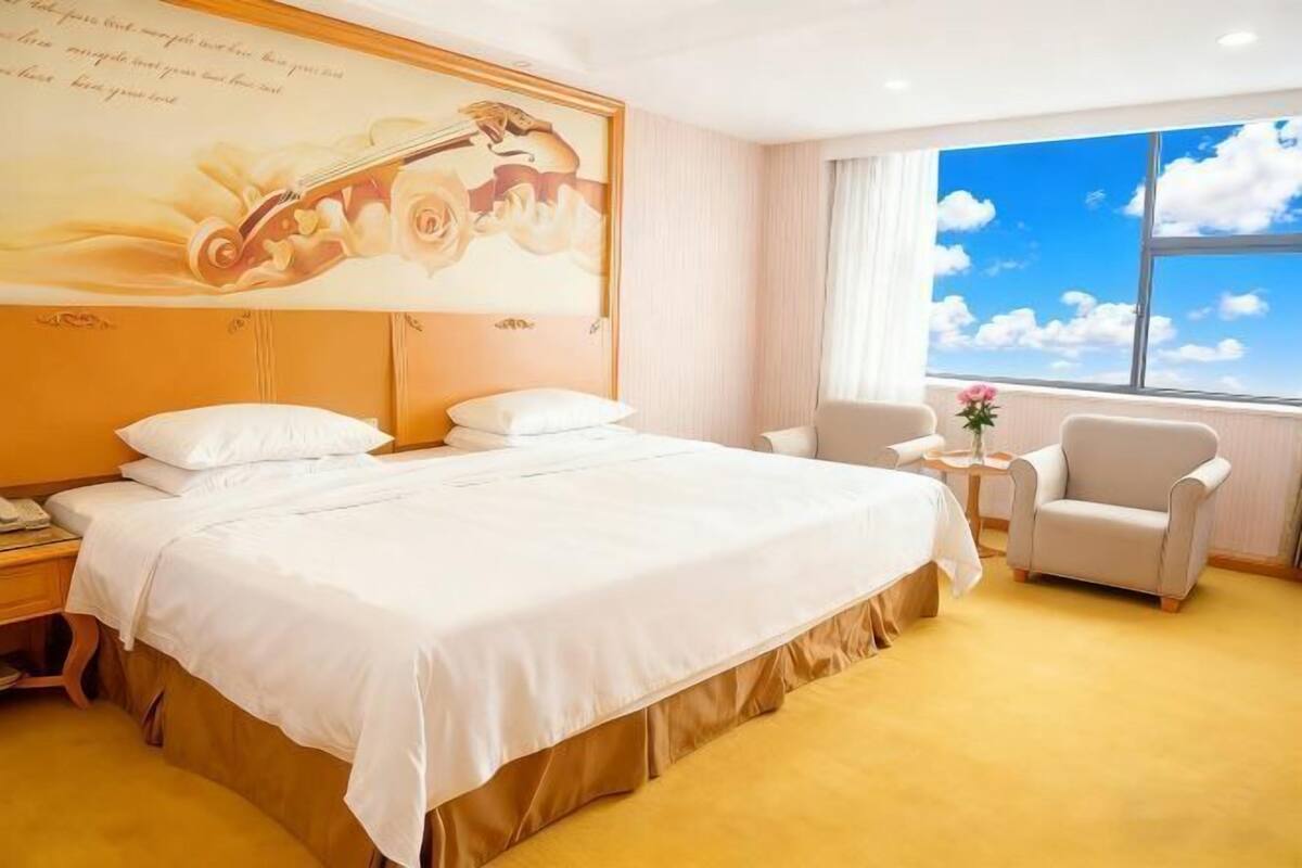 A hotel room with a large bed, a chair, a small table, and a window with a view of the sky.
