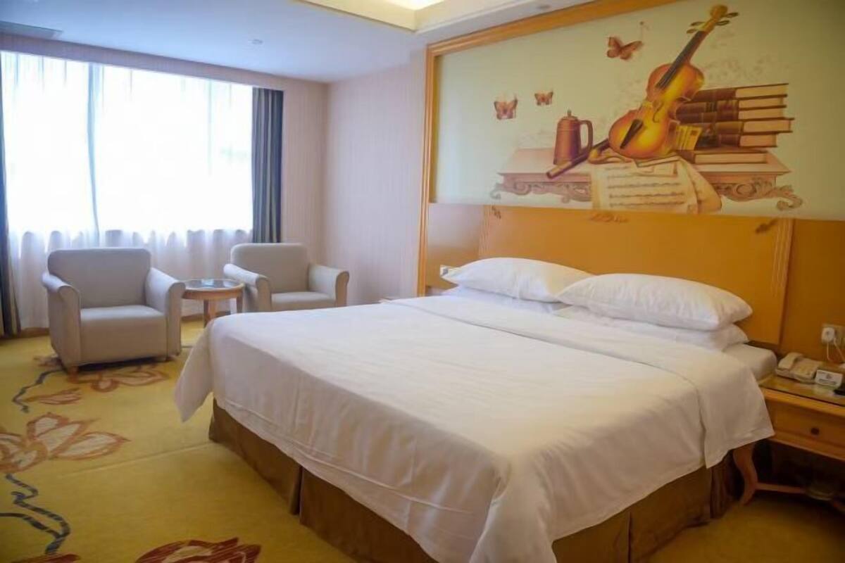 A hotel room with a large bed, two armchairs, a small table, and a wall with a musical instrument and books.
