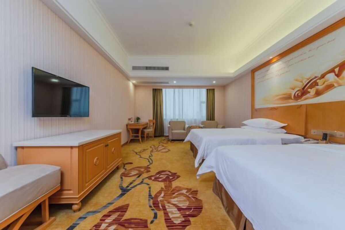 A hotel room with two beds, a TV mounted on the wall, a desk with a chair, and a floral carpet.