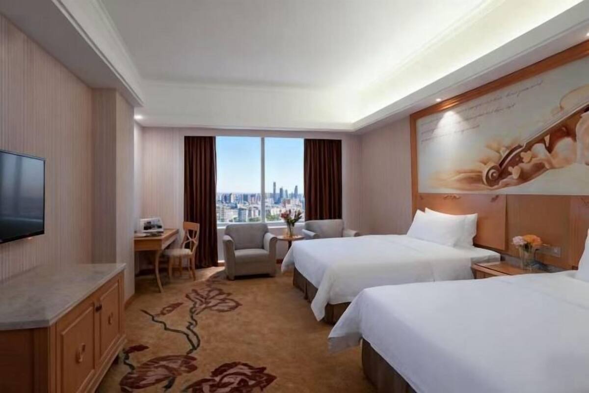 A hotel room with two beds, a desk, a chair, a TV, and a view of the city.