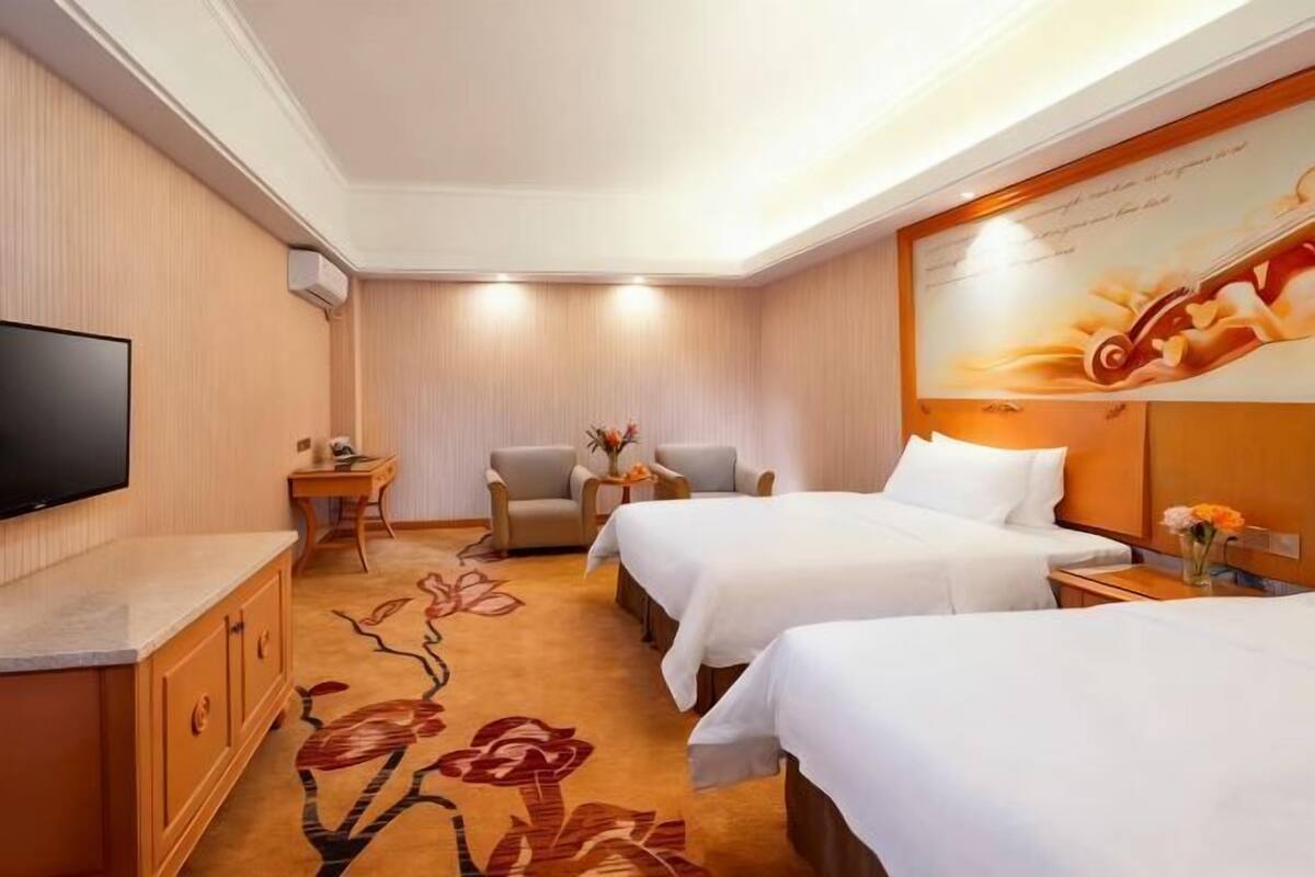 A hotel room with two beds, a TV, a desk, and a small table with flowers.