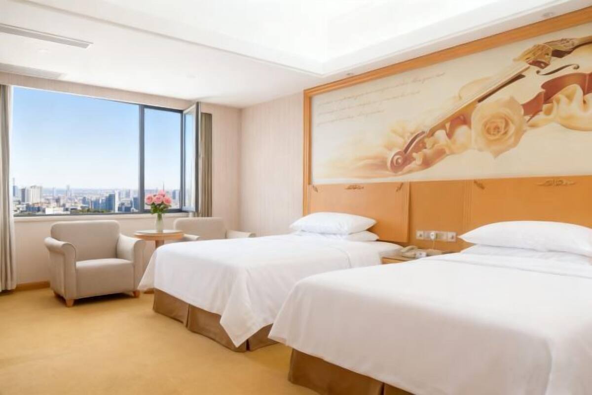 A hotel room with two beds, a sofa, a small table with a vase, and a large window with a city view.