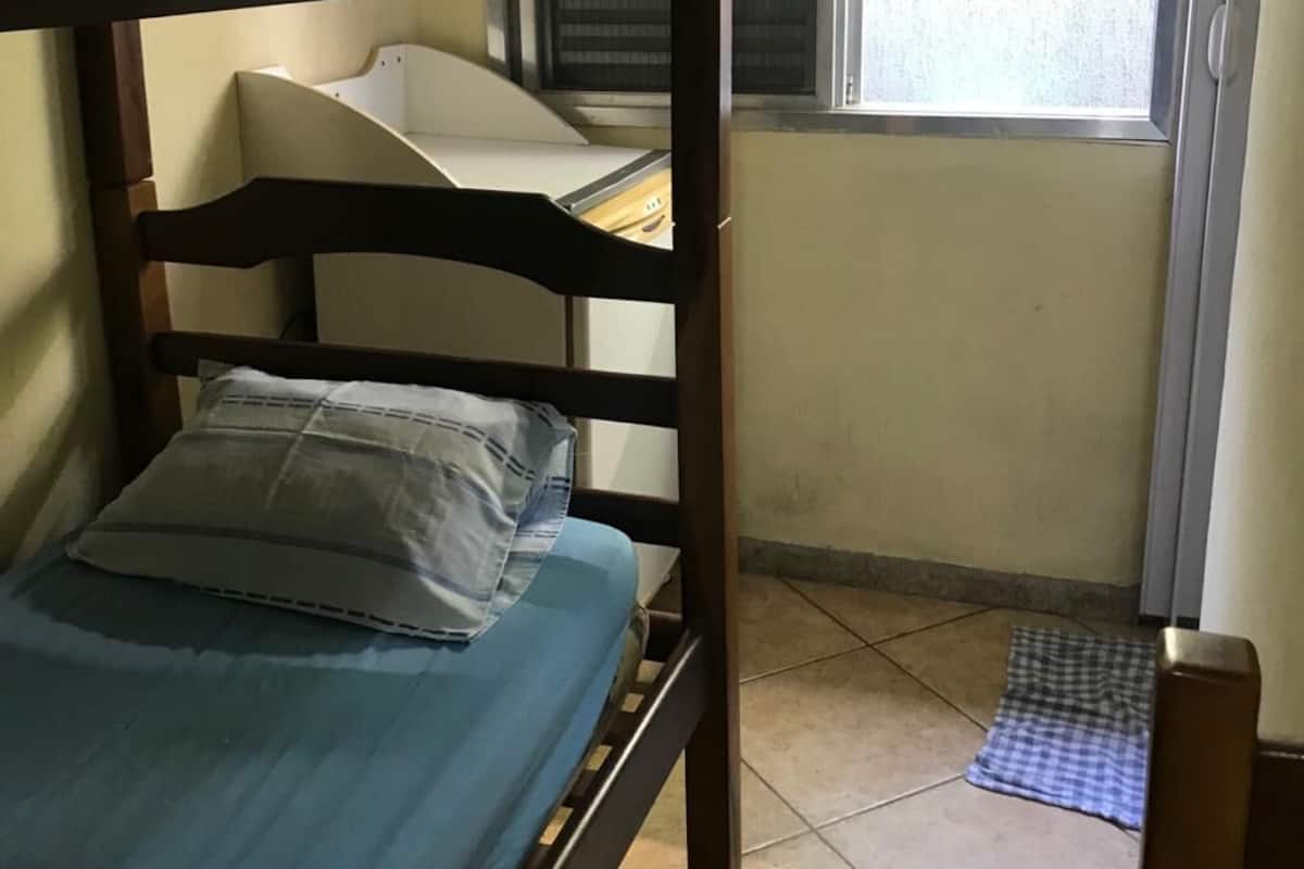 Ein Stockbett mit blauer Matratze und Kissen, ein Fenster mit Jalousien und eine karierte Matte auf dem Boden.