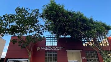 Um edifício com fachada vermelha, uma placa com a inscrição 'BALNEARIO ACAPULCO PAPELIERS DE MOGI GUACÚ' e uma entrada com portão.