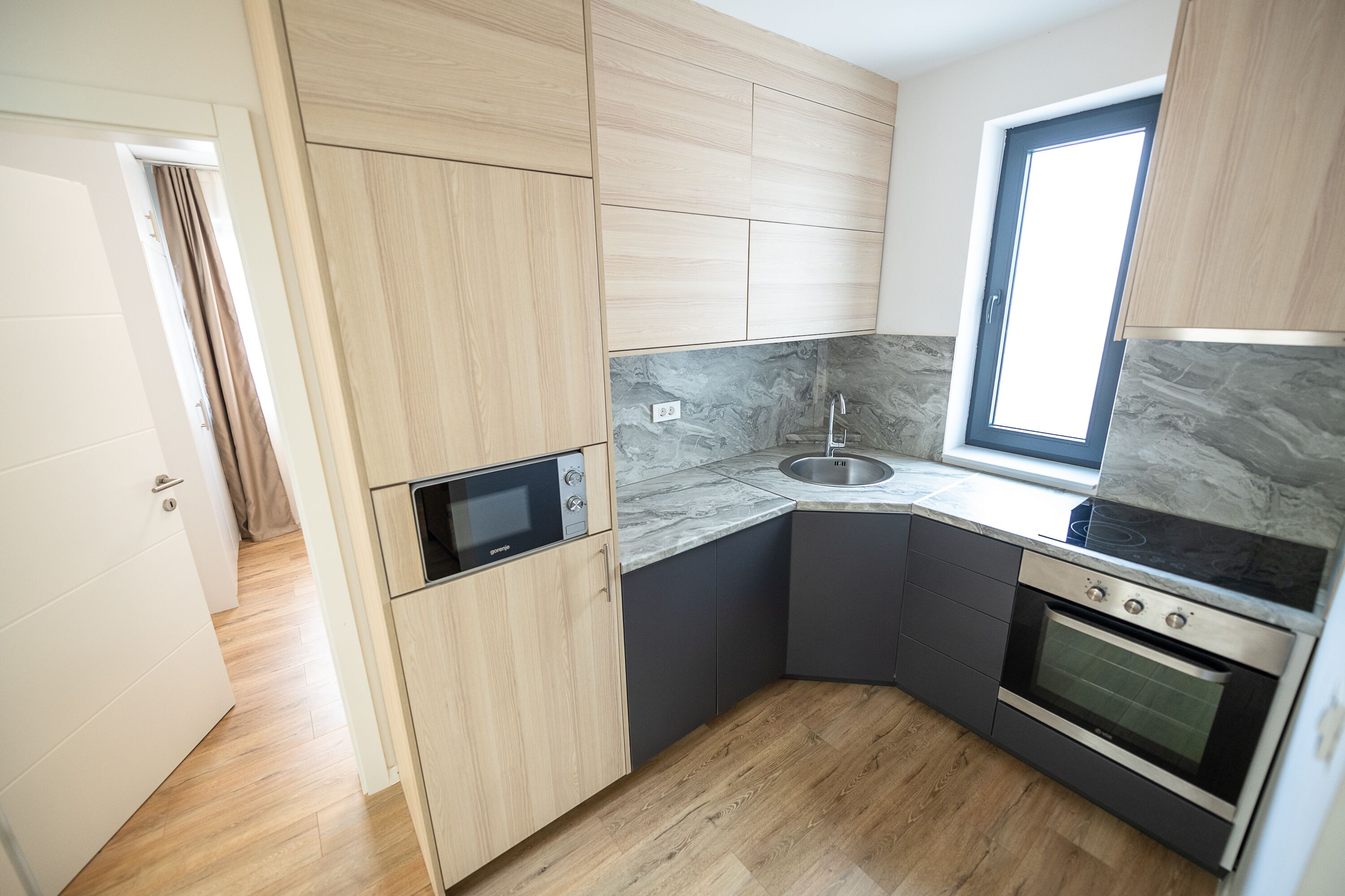 Cocina moderna con armarios de madera, horno empotrado, microondas y ventana que proporciona luz natural.