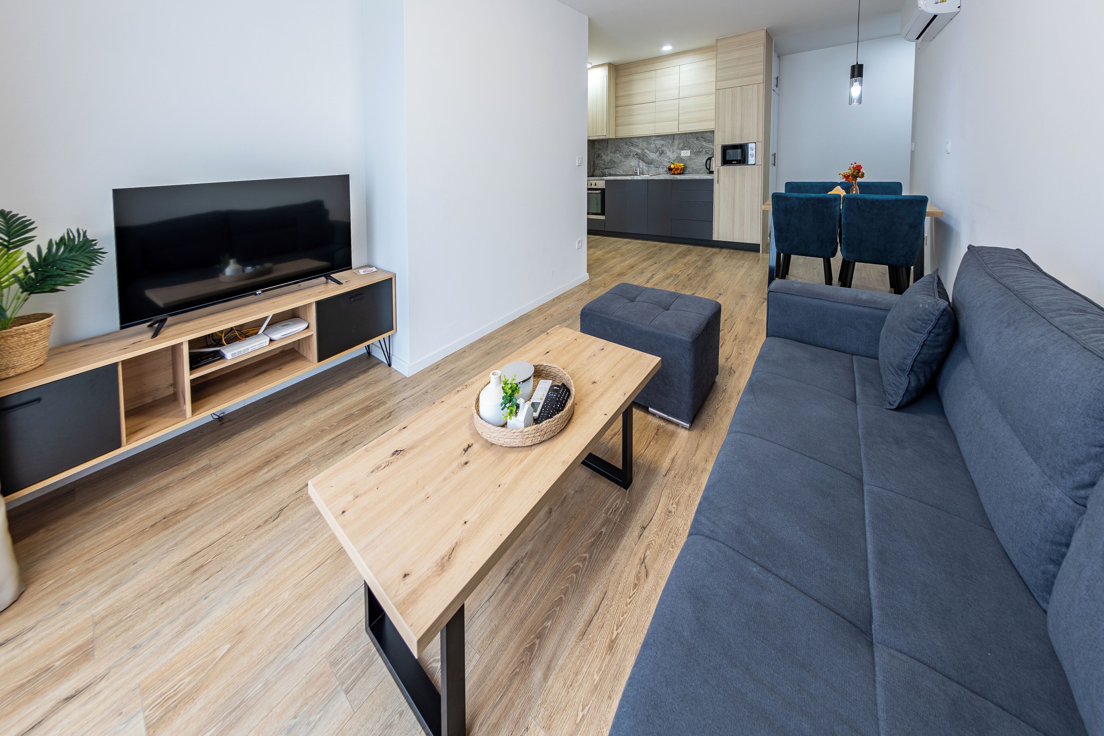 A modern living room with a flat-screen TV, wooden coffee table, grey sofa, and a kitchen in the background.