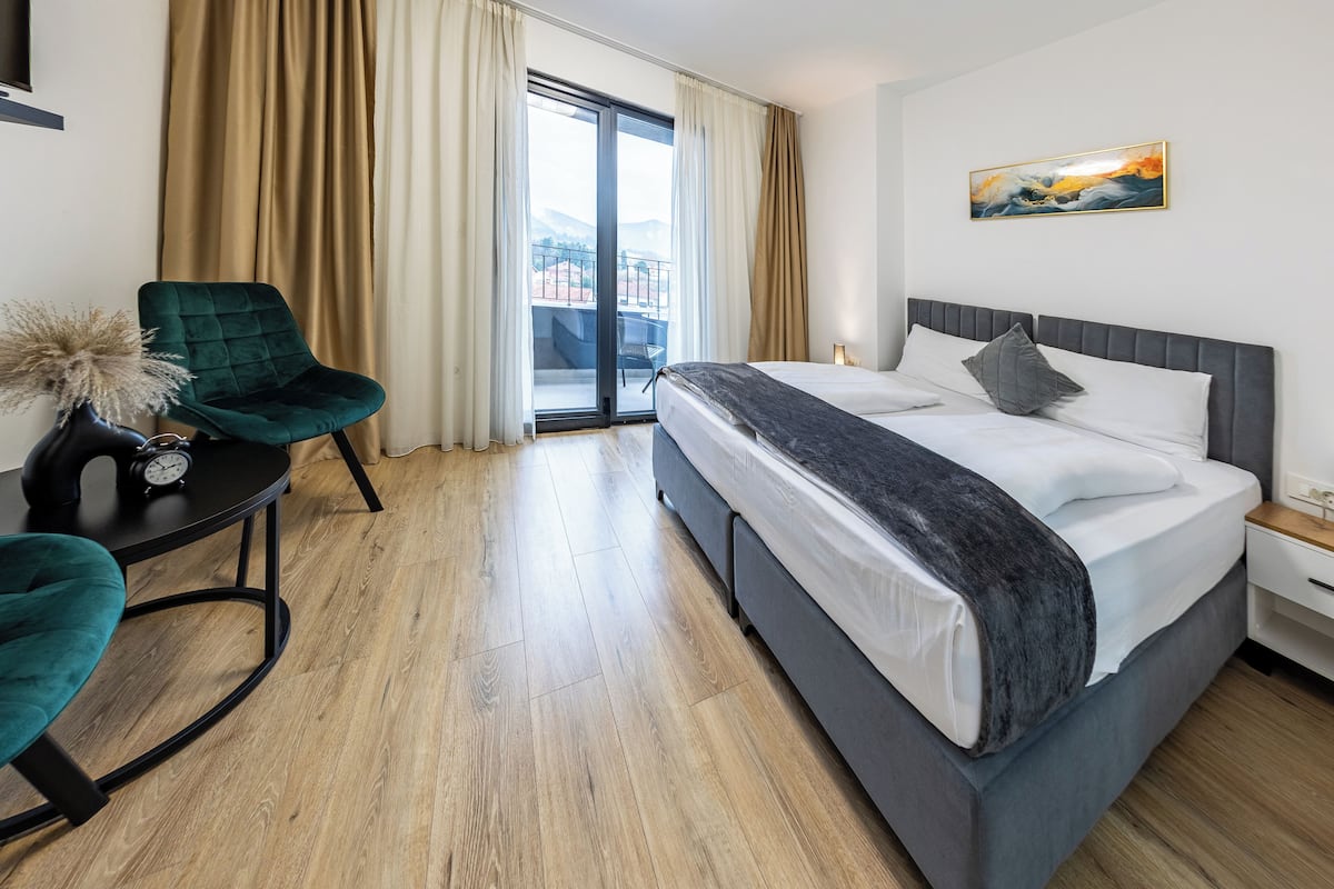 A hotel room with a bed, a chair, a table with a clock, a painting, and a view through a glass door.