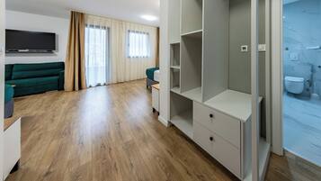Una habitación de hotel moderna con piso de madera, un sofá verde, un televisor, un armario y un baño con inodoro y ducha.