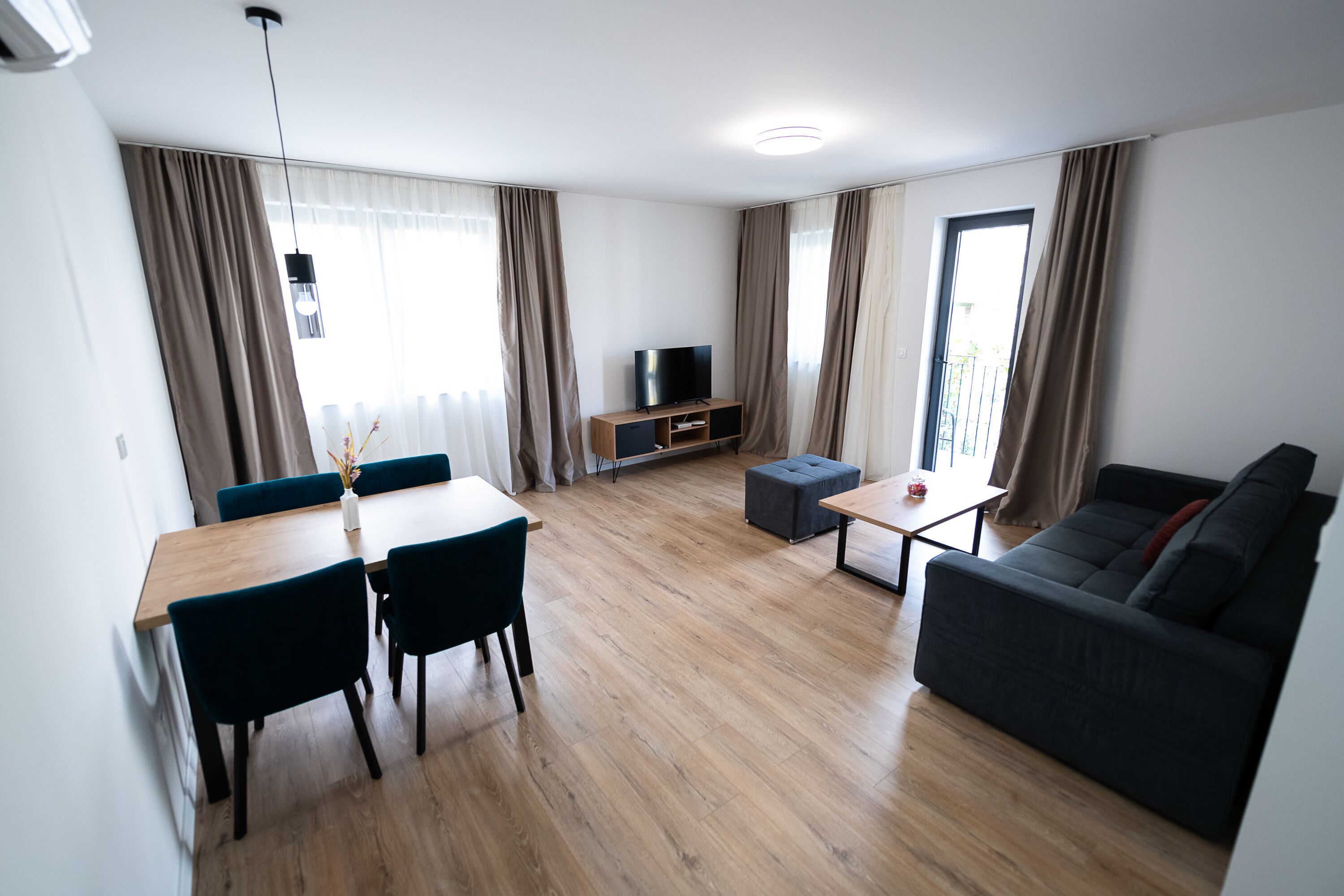 A modern living room with a wooden floor, a dark sofa, a TV on a wooden stand, a dining table with chairs, and large windows with curtains.