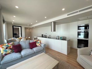 A modern living room with a grey sofa, a wooden coffee table, and a kitchen area with white cabinets and stainless steel appliances.