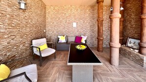 Ein Zimmer mit einer Steinwand, einer Holzbalkendecke, einem schwarzen Tisch mit einer Vase, einem grauen Sofa mit bunten Kissen und einem Stuhl mit einem gelben Kissen.
