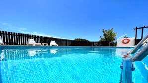 Una piscina azul cristalina con un borde de baldosas, sillas de descanso blancas y una valla de madera al fondo.
