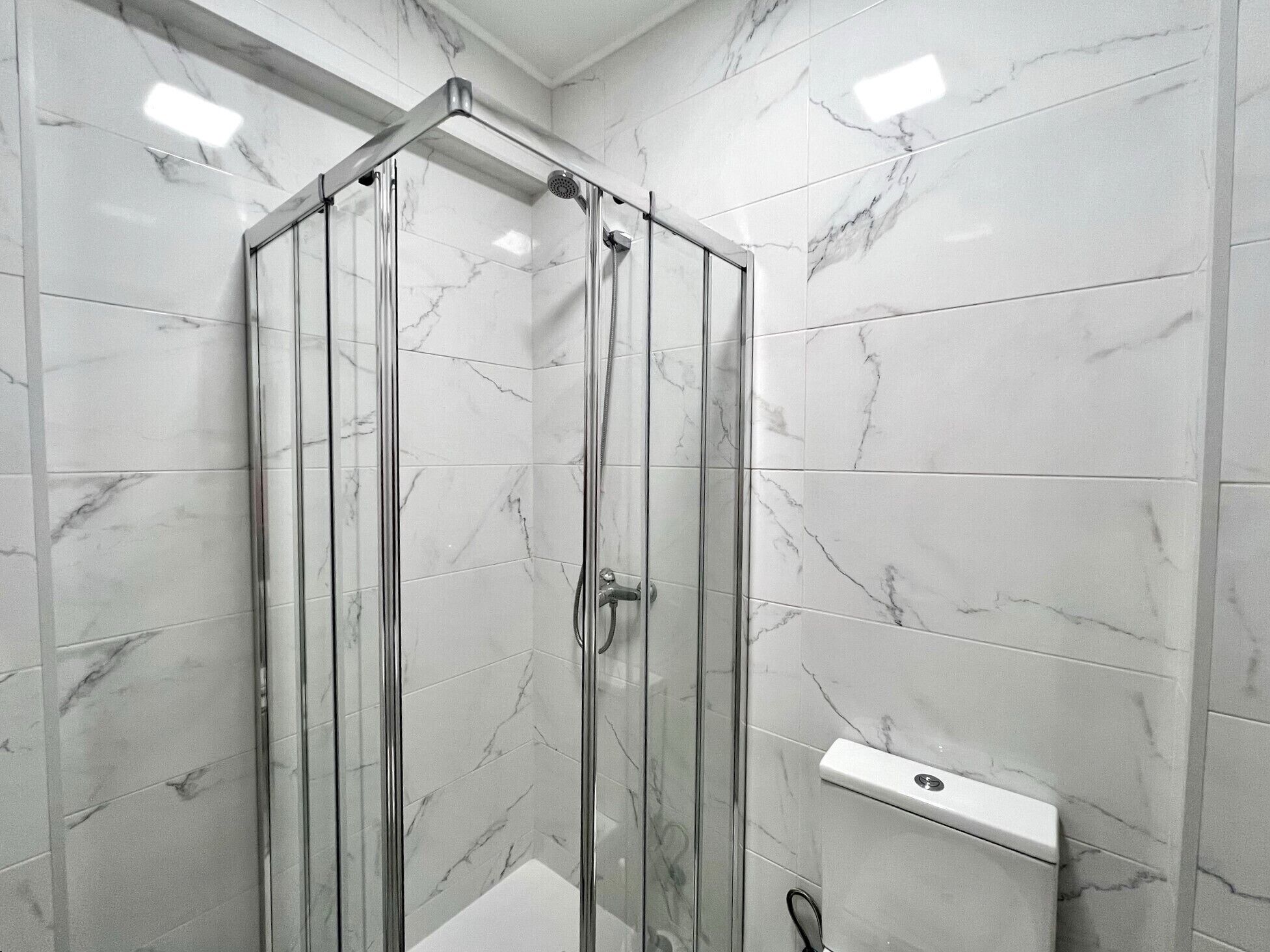 A glass-enclosed shower with a white marble wall and a white toilet.