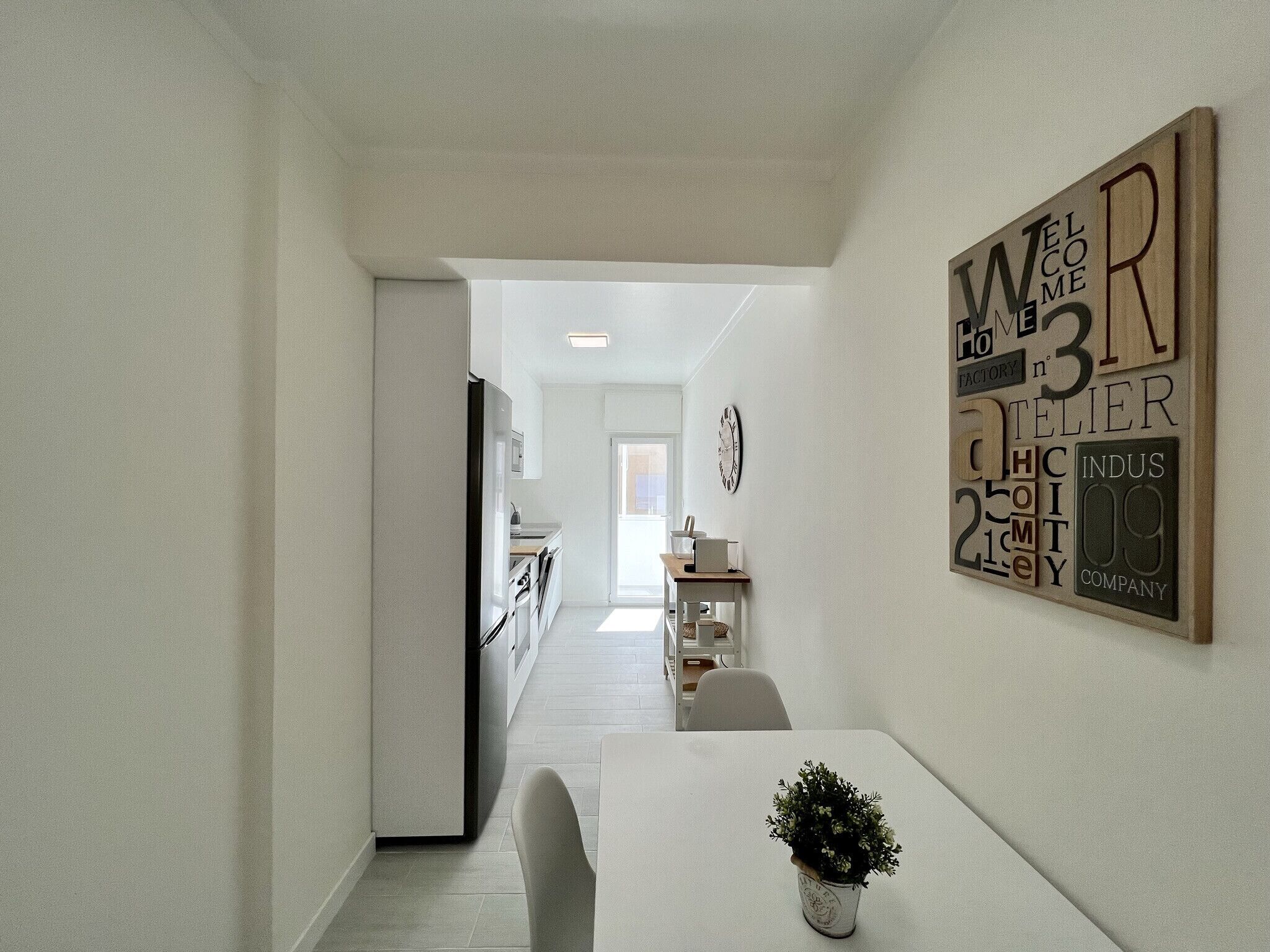 Une cuisine moderne avec des meubles blancs, un coin repas doté d’une table et de chaises blanches, et un panneau décoratif accroché au mur.
