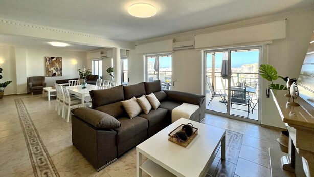 Ein modernes Wohnzimmer mit Sofa, Esstisch und Stühlen. Ein Balkon bietet Blick auf die Stadt.