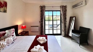 Una camera d'albergo con un letto, un comodino, una lampada rossa, una finestra con tende, una sedia e un condizionatore d'aria a muro.