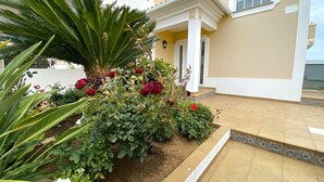 A well-maintained garden with various plants and flowers in front of a building with a tiled entrance and a balcony. - PÊRA AMAZING VILLA WITH POOL by HOMING (Pêra)