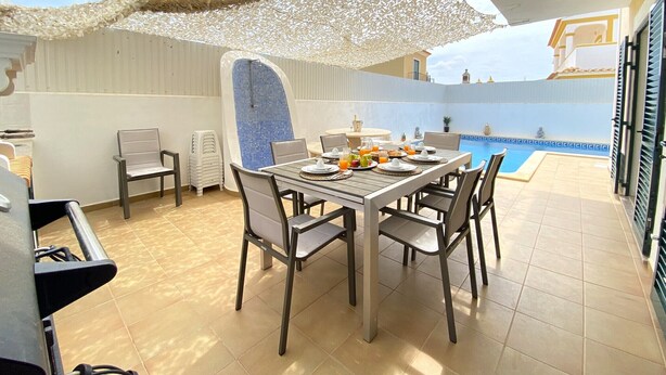 A sunlit patio with a dining table set for two, a pool in the background, and a covered seating area. - PÊRA AMAZING VILLA WITH POOL by HOMING (Pêra)