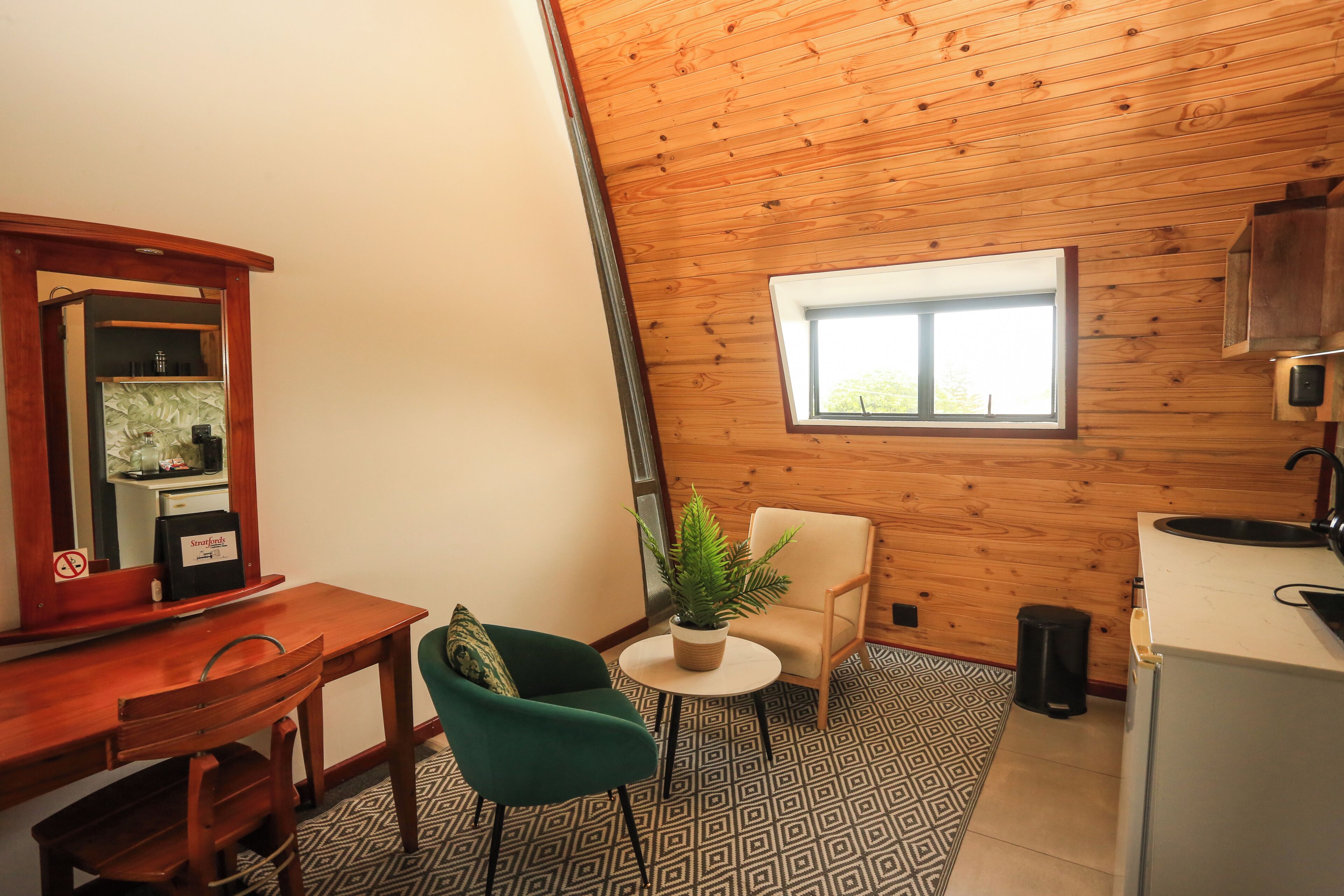 Une petite pièce agréable, avec un plafond en bois, une fenêtre, un lavabo, une chaise, une petite table sur laquelle est posée une plante, et un miroir.
