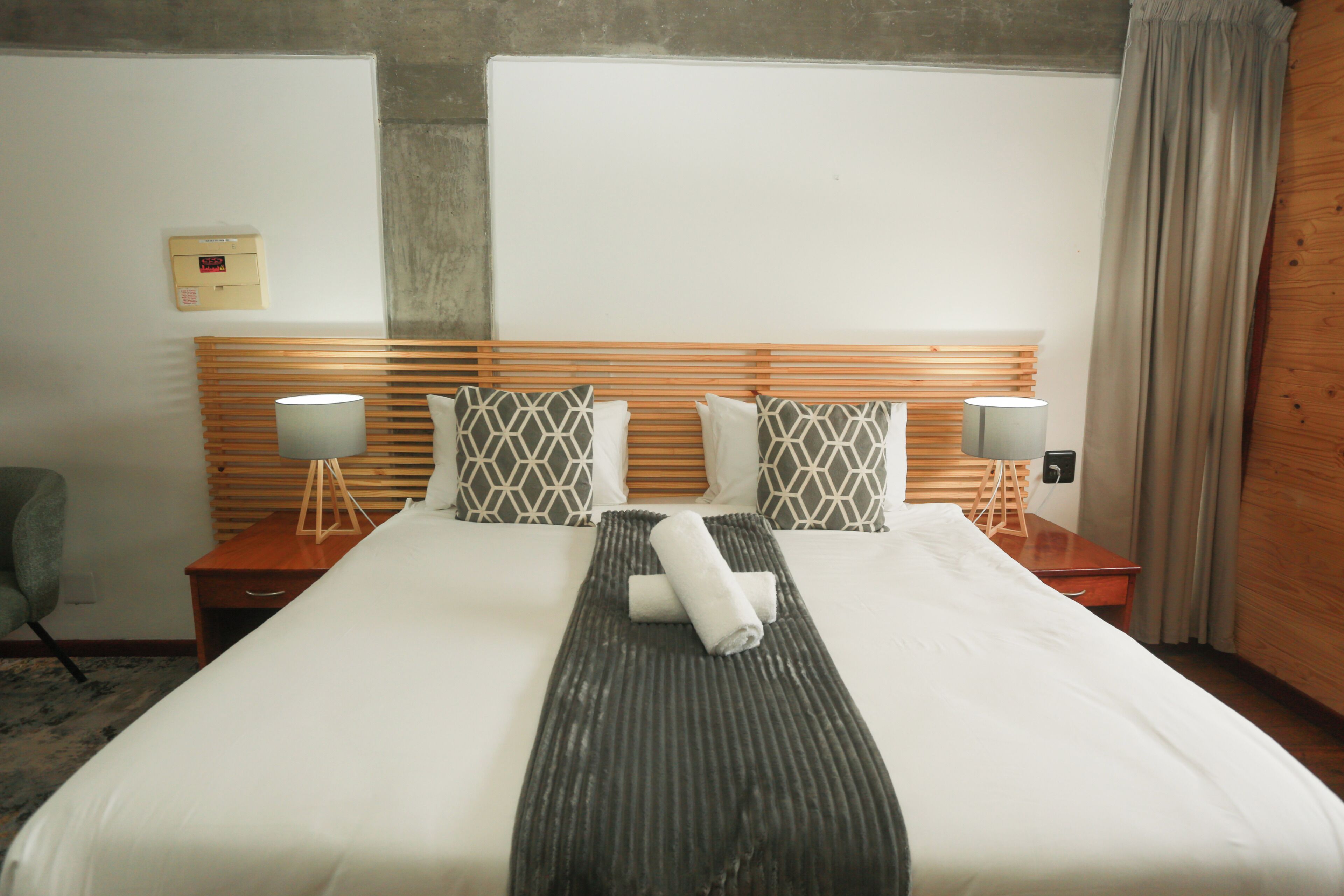 A hotel room with a large bed, wooden headboard, two bedside tables with lamps, and a grey patterned pillow.