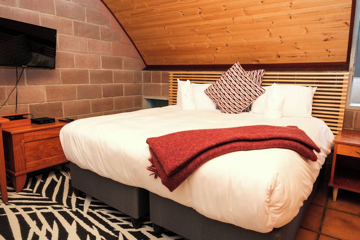 Un dormitorio con cama, cabecera de madera, televisor montado en la pared y una alfombra estampada.