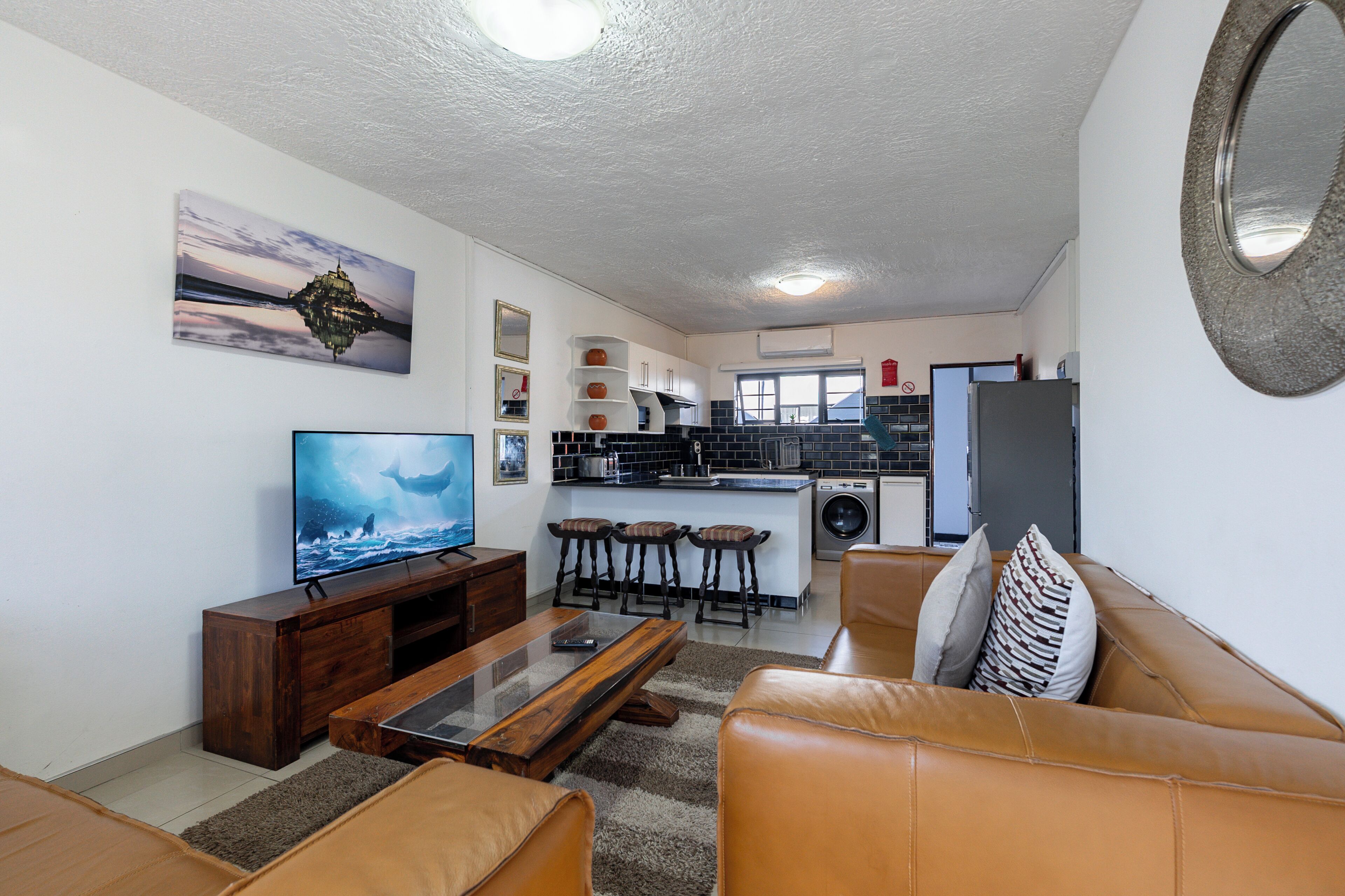 A living room with a TV, a sofa, a coffee table, and a kitchen area with a sink and stools.
