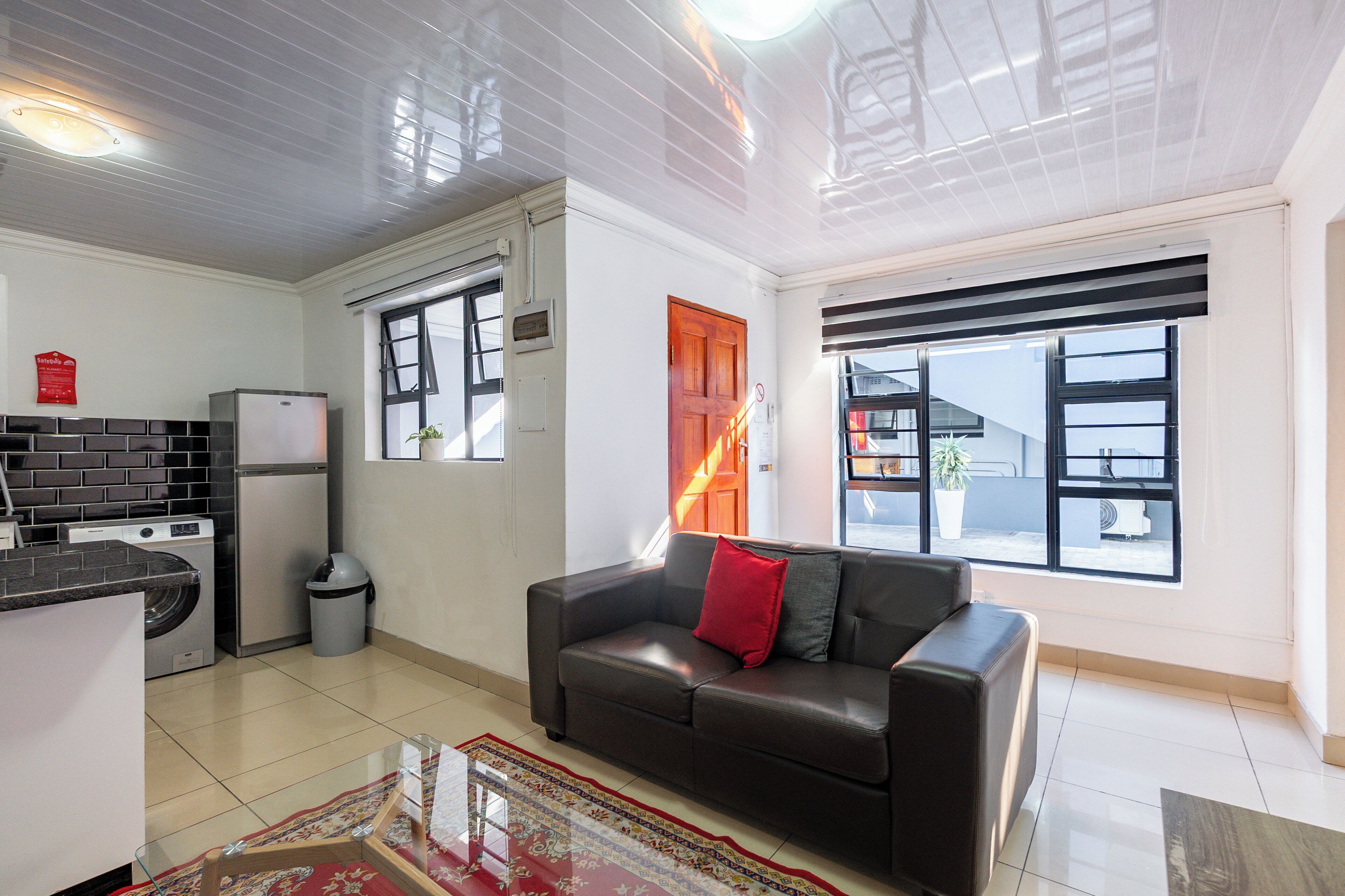 A compact living space with a kitchenette, a sofa with a red cushion, and a large window with a view.