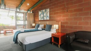 A hotel room with a large bed, a wooden headboard, a black leather sofa, a small wooden nightstand, and a large window with curtains. - Safi Self-Catering Suites (East London)