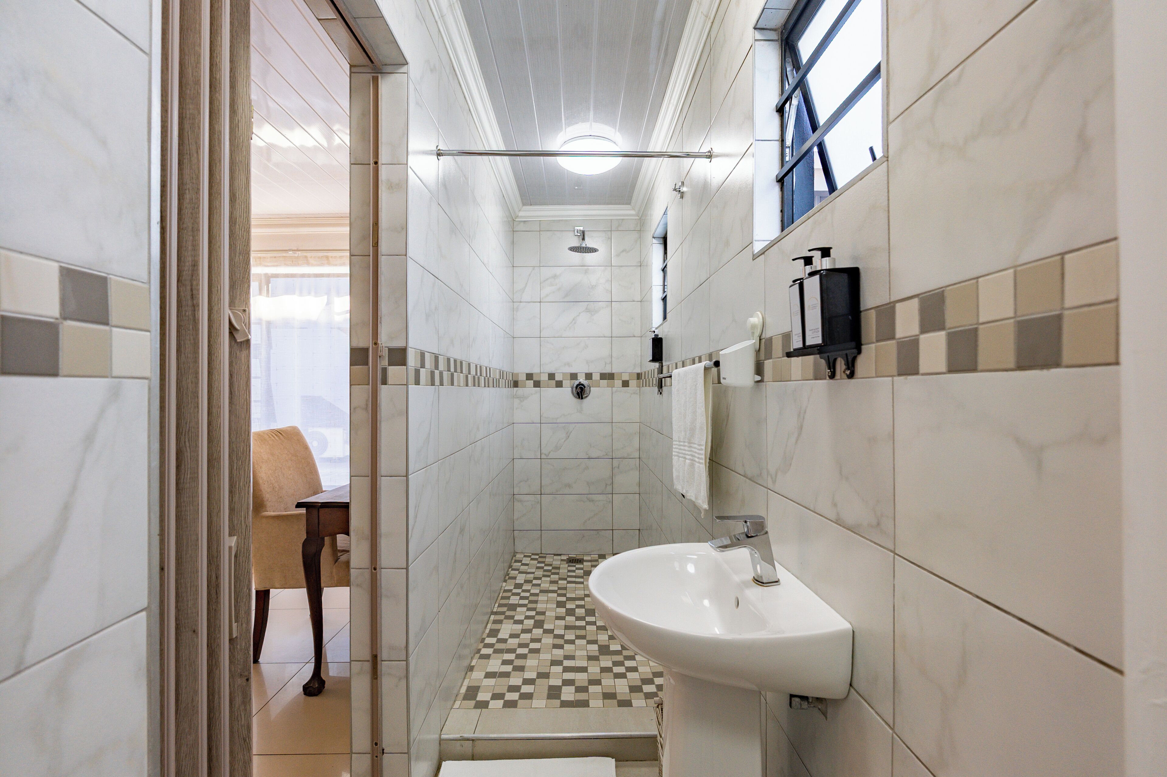 Een badkamer met douche, wastafel, stoel en raam.